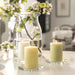 A cozy and elegant home setting featuring two lit IKEA LÖVSKOGSLUND Scented Pillar Candles with apple fragrance, placed on square glass coasters. A large glass jar behind them holds a branch with green leaves and delicate white flowers, creating a refreshing and tranquil ambiance. The soft lighting enhances the warm and inviting aesthetic.-90582065