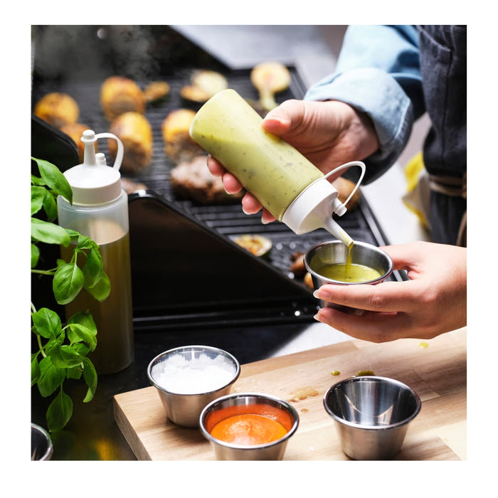 Person holding a green condiment bottle over a grill with various condiments on a wooden surface.