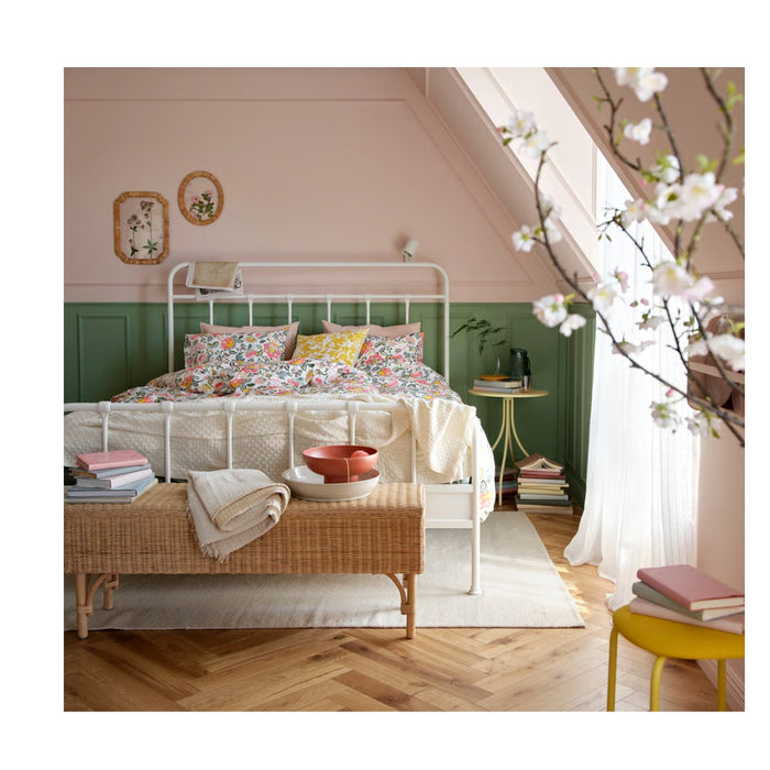 Cozy bedroom with white metal bed frame, floral-patterned duvet cover and pillows, pink and green walls, wicker bench, botanical wall art, and natural light from window.

