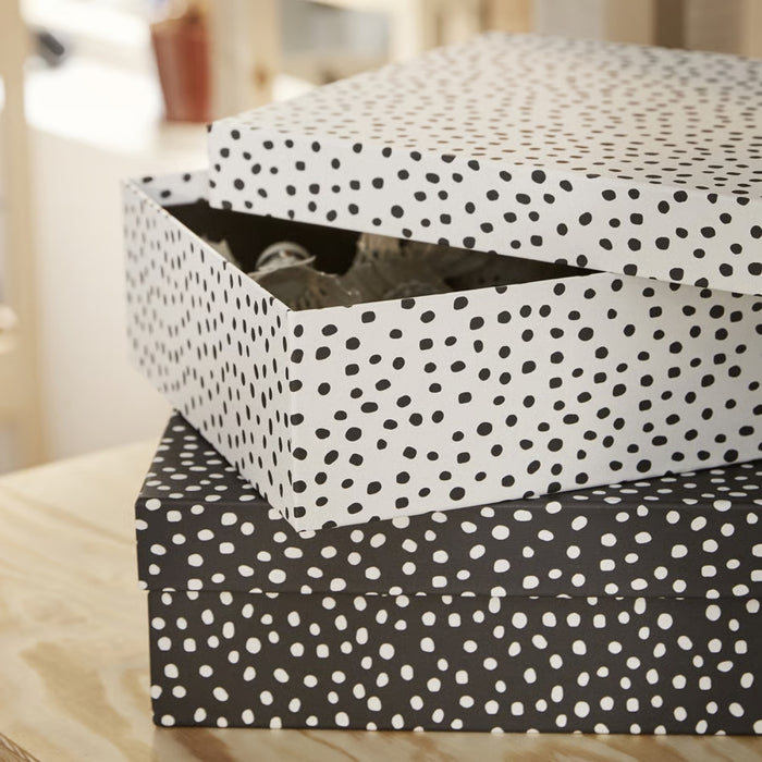 IKEA RÄTTAREN storage box set—two polka dot boxes placed on a wooden surface. Top box is white with black dots and has its lid slightly open, revealing shredded packing material inside. Bottom box is black with white dots and remains closed. Background is softly blurred, suggesting a cozy indoor setting – 90597986.