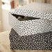 IKEA RÄTTAREN storage box set—two polka dot boxes placed on a wooden surface. Top box is white with black dots and has its lid slightly open, revealing shredded packing material inside. Bottom box is black with white dots and remains closed. Background is softly blurred, suggesting a cozy indoor setting – 90597986.