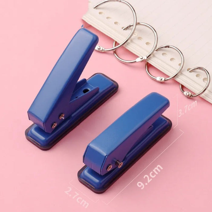 Two blue stationery hole punchers placed on a soft pink surface—one puncher measures 9.2 cm in length and 3.7 cm in width, while the smaller variant measures 2.7 cm in width. Both feature rounded handles and sturdy metal construction. A spiral-bound notebook with silver rings is partially visible in the background, suggesting practical use for organizing loose-leaf sheets – 1598448803339 13141950.