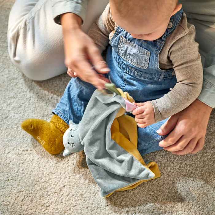Baby holding a blue and yellow cat comfort blanket while sitting on carpeted floor, playing with an adult.