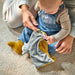 Baby holding a blue and yellow cat comfort blanket while sitting on carpeted floor, playing with an adult.