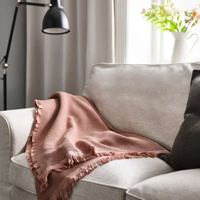 KLOTSTARR throw, pale pink, 130x170 cm – waffle-textured cotton blend with fringed edges, draped over beige sofa beside dark pillow, lamp, and floral vase in cozy living room – 50562031.