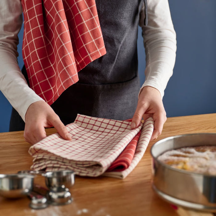 Person folding a red and white checkered towel on a wooden table with kitchen items.