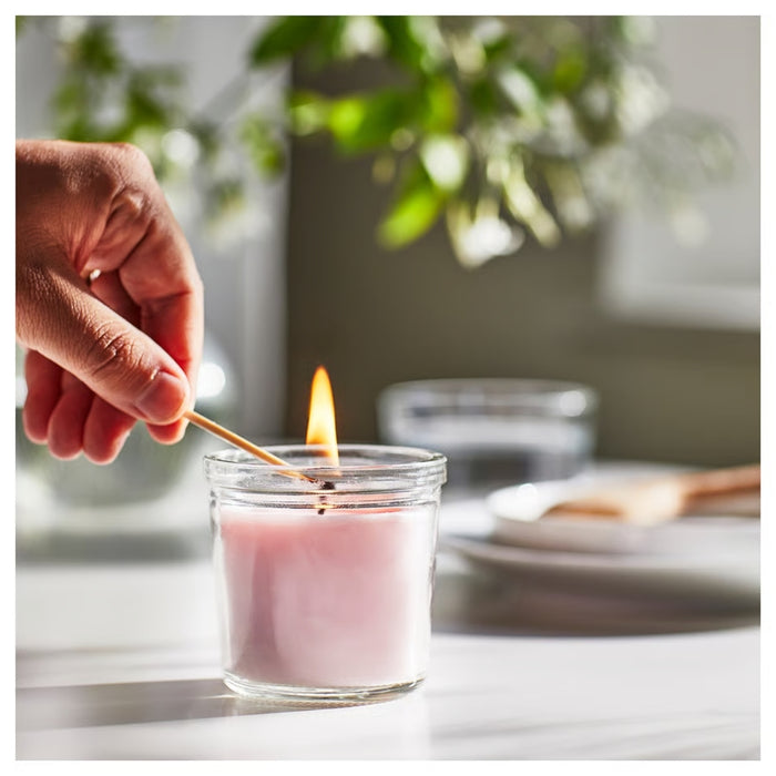 Hand lighting IKEA LUGNARE scented candle in pink glass with jasmine fragrance; 20-hour burn time; cozy home setting with soft-focus background - 00502110