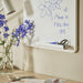 A tidy workspace featuring a whiteboard with a blue marker drawing of flowers and the message “Have a nice day” with hearts. Below, a tray holds scissors and a marker, while a desk displays a vase of purple and white flowers, papers, and a white desk lamp.