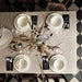 Dining table setup for four with a beige tablecloth, white plates, white bowls, black napkins tied with twine, silverware, wine glasses, and a vase with dried flowers—elegant setting for stylish dining and home decor.