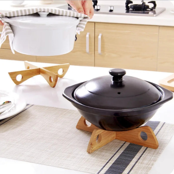 Kitchen setting featuring two cooking pots placed on wooden triangular trivets with circular cutouts—one pot is white with side handles and is being lifted by a person using a striped kitchen towel; the second pot is black with a lid and knob, resting nearby. Both trivets help protect the countertop from heat. Background includes a stovetop and cabinetry, framing a cozy and functional culinary scene – 562152 12826862.