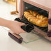 Hand using a black heat-resistant oven mitt to remove a tray of golden brown bread from a compact pink electric oven. The oven door is open, revealing a glass window and two silver control knobs on the side. The scene captures a moment of safe, home baking—emphasizing kitchen safety and the mitt’s insulation function.-1598452898401 13017586