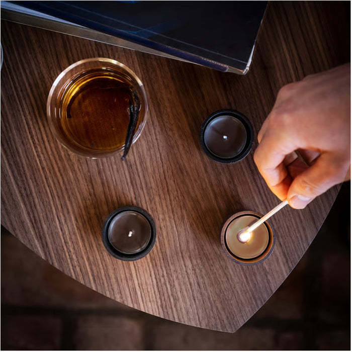 IKEA SILKESTRÄD scented tealights in black holders with leather and vanilla fragrance; hand lighting candle beside warm drink and book on wooden table – 3.5-hour burn time, 30-pack - 20570551