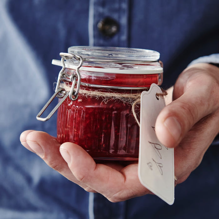 Hand holding a jar of red jam with a tag, against a blurred blue background