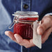 Hand holding a jar of red jam with a tag, against a blurred blue background