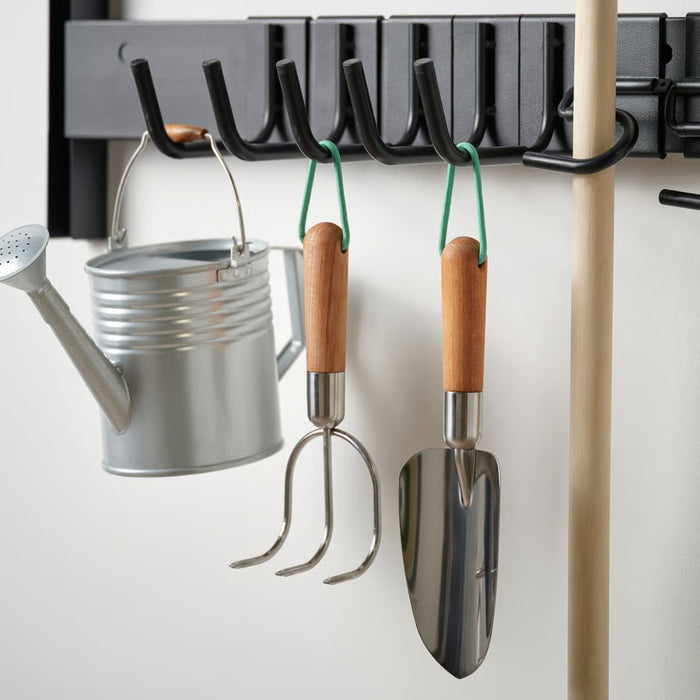 Wall-mounted rack with stainless steel gardening tools including a trowel and cultivator, both with birch handles and green loops, neatly organized alongside a watering can.