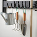 Wall-mounted rack with stainless steel gardening tools including a trowel and cultivator, both with birch handles and green loops, neatly organized alongside a watering can.