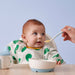 Baby being fed with a spoon by an adult against a light blue background