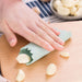 Person using a garlic peeler on cloves of garlic with a wooden board and striped cloth in the background.
