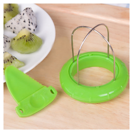 A green plastic fruit slicer with a metal wire loop and a matching green plastic blade, shown on a light wood surface next to a plate of sliced kiwi and dragon fruit.