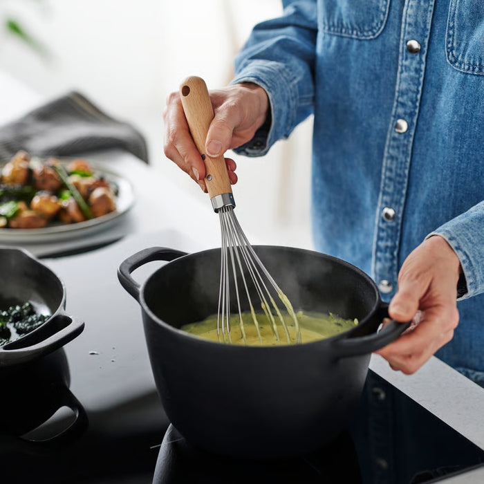 A person in a denim shirt is whisking a steaming mixture in a black pot on a stovetop, using the IKEA VARDAGEN balloon whisk. The scene captures an active cooking moment with visible steam, suggesting the food is being prepared hot. In the background, there's a plate of roasted vegetables and greens, while another pot with dark green contents sits nearby. The warm, rustic kitchen setup emphasizes the whisk’s professional quality and versatility in real culinary use -00581485.