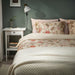 Bedroom with floral bedding and pillows, nightstand with books and lamp.
