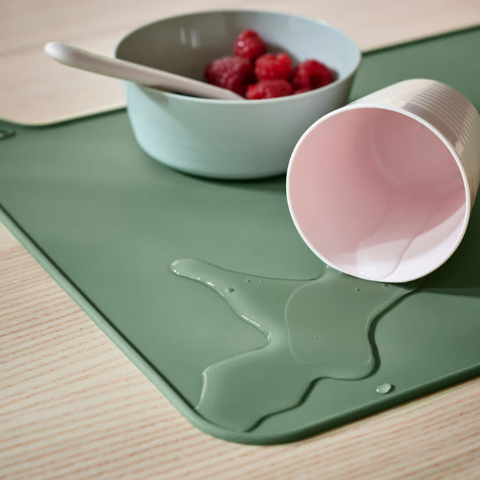 Close-up of light green silicone placemat with tipped-over cup and spilled liquid contained on its surface.

