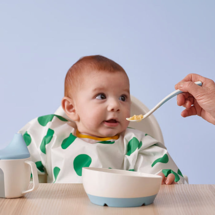 Baby in white bib with green polka dots seated in high chair; being fed with IKEA BÖRJA white/blue spoon from matching bowl – sippy cup placed on table, set ideal for safe and ergonomic infant feeding - 70596817