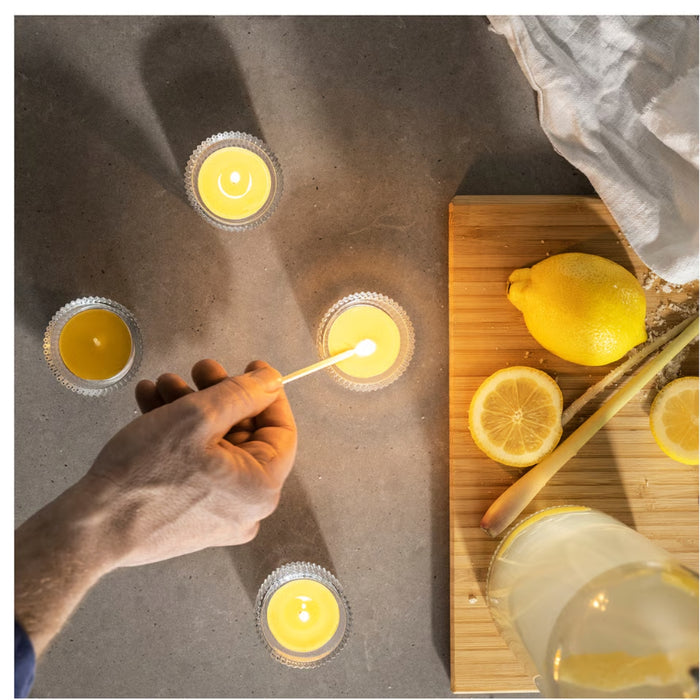 IKEA BLODHÄGG scented tealight – lemon & lemongrass/yellow, hand lighting candle beside lemons and lemonade - 30571507