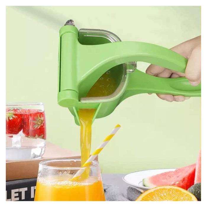 A green manual citrus juicer being squeezed by a hand, with orange juice pouring into a glass with a striped straw. Fruits like watermelon, orange, and strawberries are visible around it on a tabletop against a pale green background.