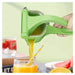 A green manual citrus juicer being squeezed by a hand, with orange juice pouring into a glass with a striped straw. Fruits like watermelon, orange, and strawberries are visible around it on a tabletop against a pale green background.