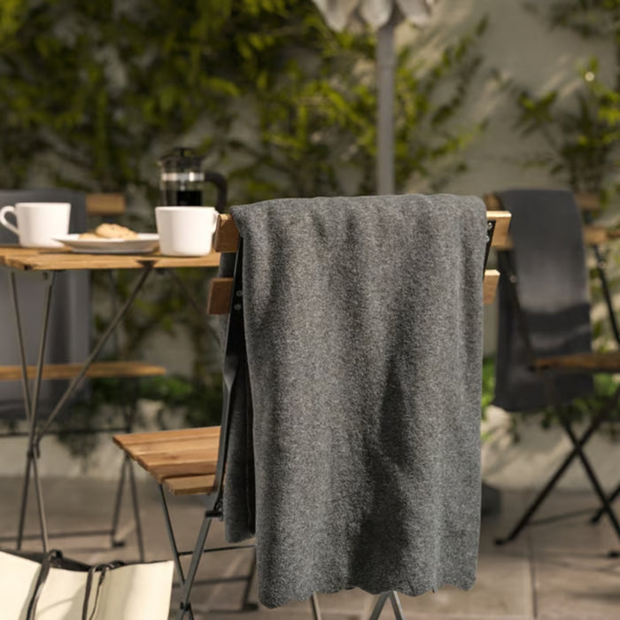 DAGGKÅPOR throw, dark grey, 130x170 cm. Draped over outdoor chair in patio setting with coffee mugs and French press on table – adds cosy touch to garden mornings – 50598643.