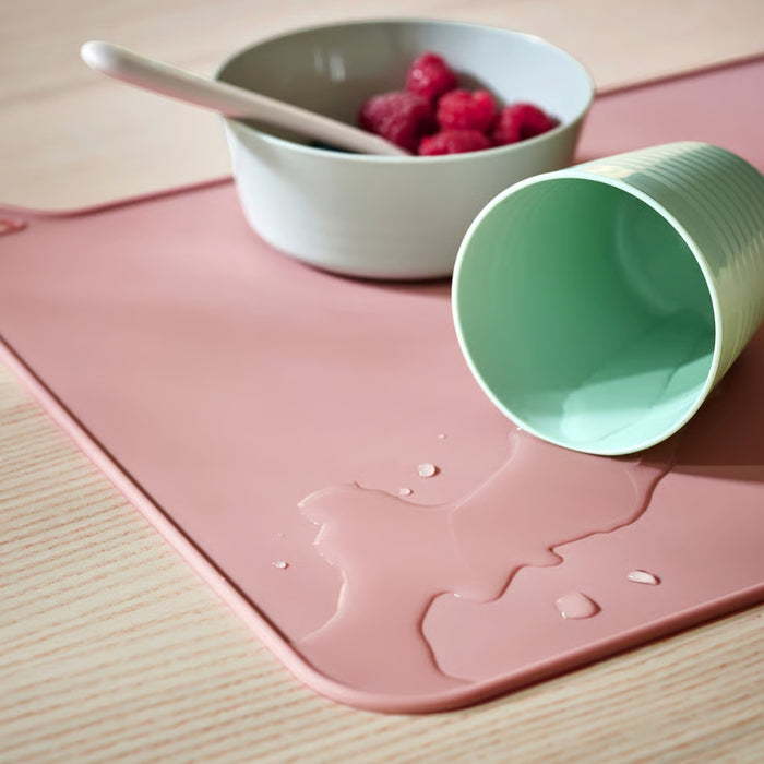 Close-up of pink silicone placemat with tipped-over mint green cup and spilled liquid contained on its surface.

