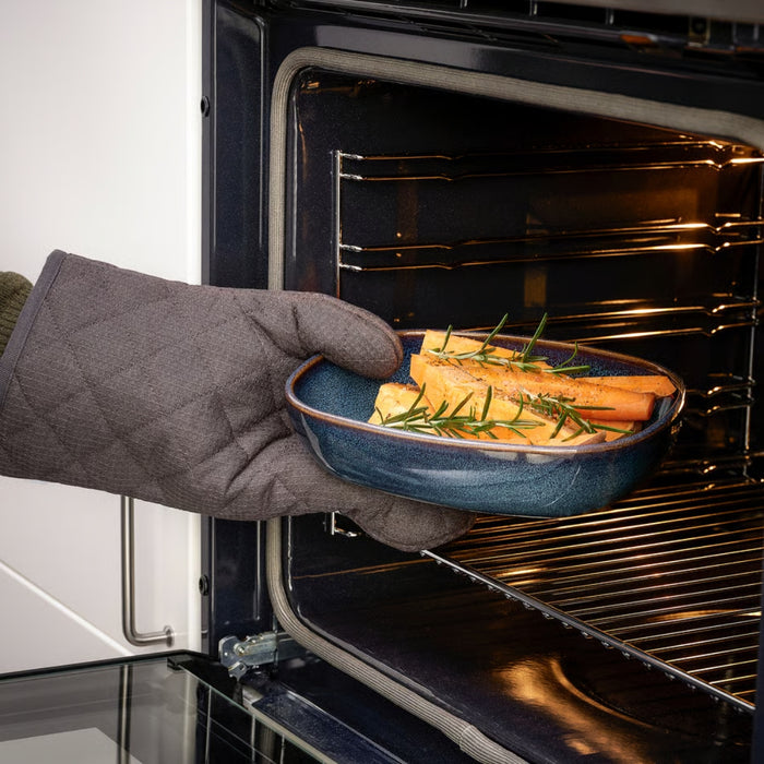 A hand wearing a gray oven mitt is seen placing or retrieving a blue ceramic dish from an oven. The dish, part of the IKEA GLADELIG set, contains golden-baked vegetables—likely potatoes and carrots—garnished with sprigs of rosemary. Warm light from the oven highlights the dish’s rich blue tone and glossy finish, conveying both utility and visual appeal in a cozy kitchen setting -20531079.