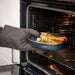 A hand wearing a gray oven mitt is seen placing or retrieving a blue ceramic dish from an oven. The dish, part of the IKEA GLADELIG set, contains golden-baked vegetables—likely potatoes and carrots—garnished with sprigs of rosemary. Warm light from the oven highlights the dish’s rich blue tone and glossy finish, conveying both utility and visual appeal in a cozy kitchen setting -20531079.