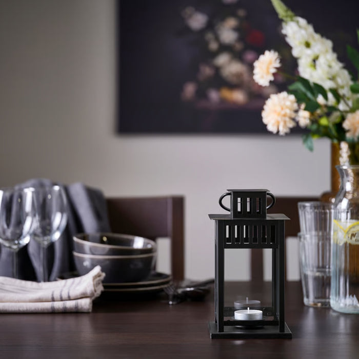 Black lantern with a candle on a dining table with floral arrangement and glasses in the background
