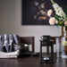 Black lantern with a candle on a dining table with floral arrangement and glasses in the background