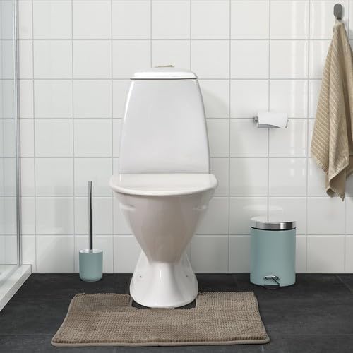 Bathroom scene featuring an IKEA TOFTBO pedestal mat in beige positioned around the base of a white toilet—set against a white tiled wall, the space includes coordinated accessories: light blue toilet brush holder to the left, a beige towel hanging on a hook, a wall-mounted toilet paper holder, and a light blue trash can with silver lid. The mat rests on a dark flooring surface, providing warmth, comfort, and water absorption in a clean, modern layout –50610268