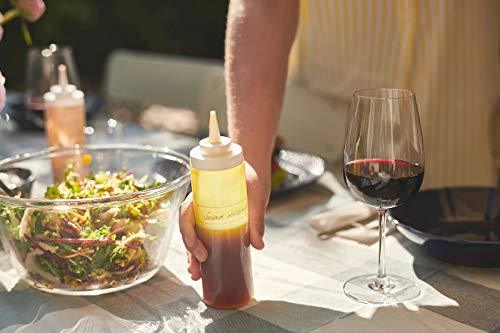 Person holding a bottle of La Tourangelle balsamic vinaigrette over a salad with a glass of red wine on a table.