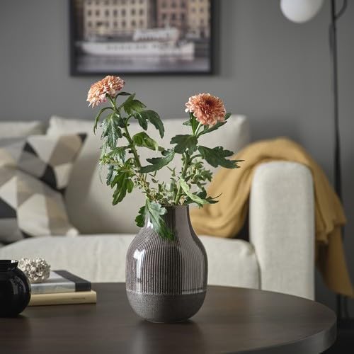 IKEA GRADVIS Vase, dark grey, 21 cm (8¼"), displayed on a round wooden coffee table in a cozy living room setting. The ribbed ceramic vase holds two pink-orange flowers with green foliage, adding a delicate touch to the space. In the background, a beige sofa with a geometric cushion and a yellow throw creates a warm atmosphere, complemented by a framed cityscape on the wall. A black decorative sculpture and a book placed beside the vase complete the stylish setup.