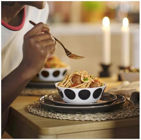 Dining scene featuring a person enjoying spaghetti with meatballs from an IKEA OFTAST bowl—white bowl adorned with black polka dots is placed on a black plate with a woven placemat underneath.-10608267