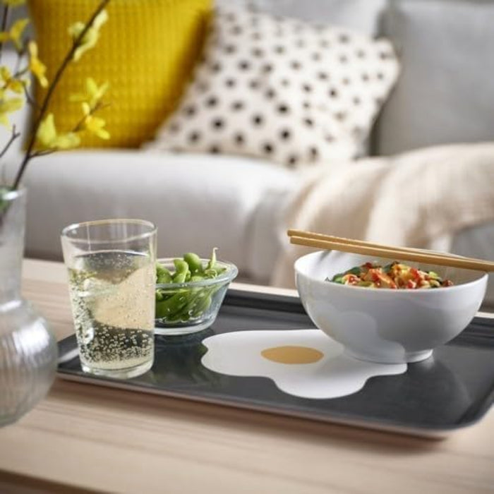 Lifestyle scene featuring a person holding the IKEA KEJSARFISK tray with a bold fried egg graphic in the center—tray supports a white bowl of noodle soup, a small glass dish of edamame, and a pair of wooden chopsticks resting across the tray. – 50607690.