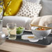 Lifestyle scene featuring a person holding the IKEA KEJSARFISK tray with a bold fried egg graphic in the center—tray supports a white bowl of noodle soup, a small glass dish of edamame, and a pair of wooden chopsticks resting across the tray. – 50607690.