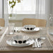Dining table setup featuring two coordinated place settings with IKEA OFTAST bowls—each white bowl displays a black circular dot pattern around the rim and is paired with matching white plates and silver cutlery placed on beige napkins.10608267