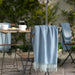 Outdoor dining scene featuring a wooden table and chairs set against lush greenery and a stone wall backdrop. On the table are two white cups, a ceramic plate with food, and a glass coffee pot. A light blue IKEA HOLMVI throw is draped casually over the back of one wooden chair, adding a touch of cozy texture and color to the alfresco setup – 80604685.