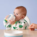 : Lifestyle scene featuring a young child seated at a table, holding and drinking from the IKEA BÖRJA training beaker in white and blue—child wears a bib with green polka dots and yellow cuffs, adding playful vibrancy. A matching white and blue plate sits on the table, and an adult hand rests nearby, subtly emphasizing caregiver supervision. The soft blue background enhances the calm, nurturing atmosphere of mealtime training.-50596818