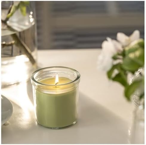 Lit green candle in glass placed beside vase with leaves and white flowers – 50582053