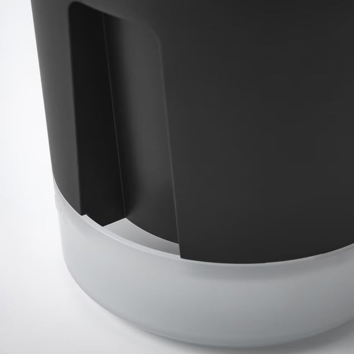 IKEA VÅRDTRÄD self-watering plant pot—transparent with anthracite base, shown on a tabletop near green foliage and a watering can; vertical cut-out detail offers a glimpse of the inner reservoir design – 80570869.