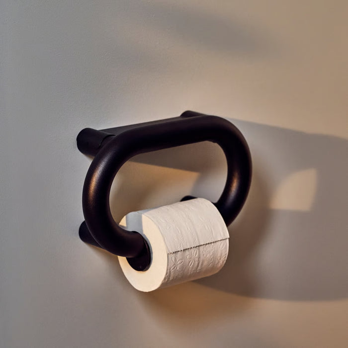 Black metal toilet paper holder with a roll of toilet paper on a plain wall.