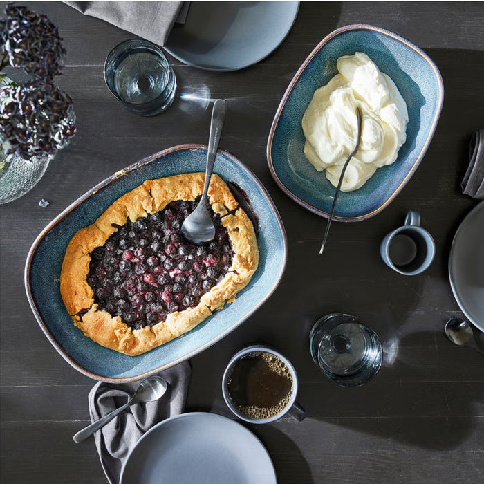 A beautifully styled table setting featuring an IKEA GLADELIG blue oven dish filled with a rustic berry galette. The dessert’s golden crust and juicy dark berry center are complemented by a second matching dish holding whipped cream. Additional table elements include a coffee mug, glass of water, small bowl, napkin with spoon, and a vase of deep purple flowers, creating a cozy yet elegant ambiance perfect for a homemade treat or brunch gathering -20531079.