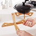 Hands assembling a cross-shaped wooden trivet in a kitchen setting—two interlocking bamboo slats with rounded edges and circular cutouts form an “X” base; a black pot with lid rests on a similar assembled trivet in the background. Kitchen counter includes white ceramic containers and a stove, highlighting the trivet’s use for heat protection during cooking – 562152 12826862.